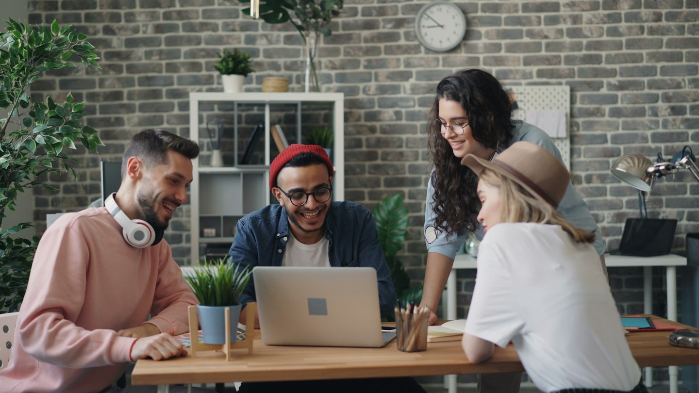 Quattro giovani professionisti sorridono e collaborano attorno a un laptop su una scrivania.