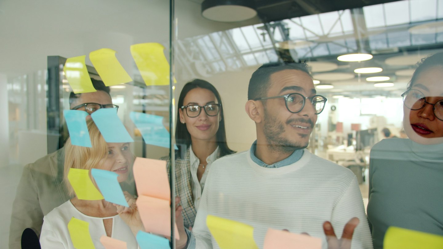 Team eterogeneo che discute idee con post-it su vetro.